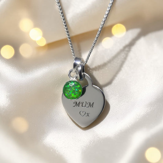 Silver heart-shaped pendant with green gemstone and 'MUM' engraving on a soft fabric background