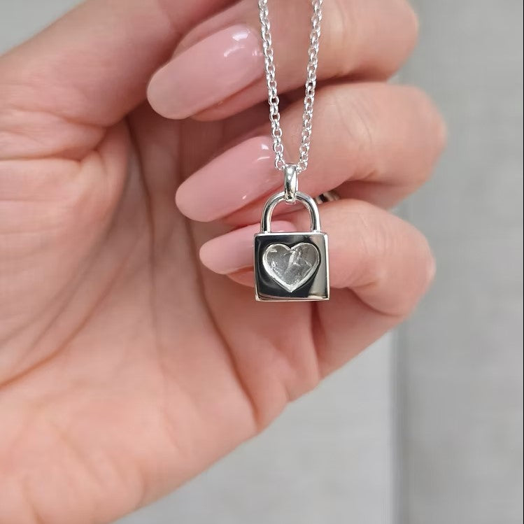 Silver padlock-shaped pendant with a heart design on a chain held in a hand.