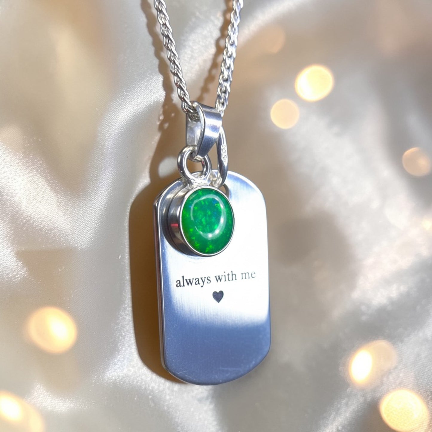 Silver pendant with green gemstone and 'always with me' engraving on a soft fabric background